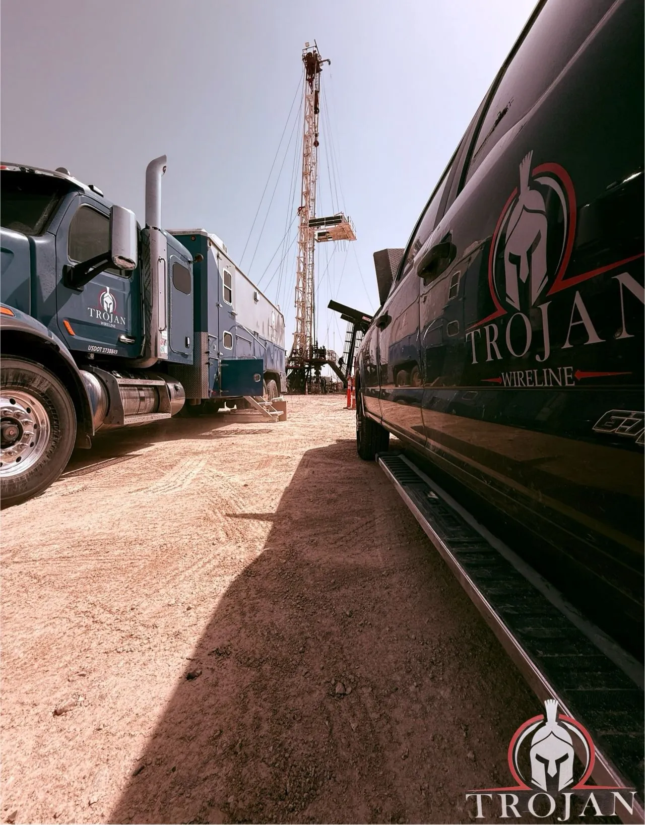 Low-angle view of two Trojan Wireline trucks parked at a drilling rig under a clear sky.
